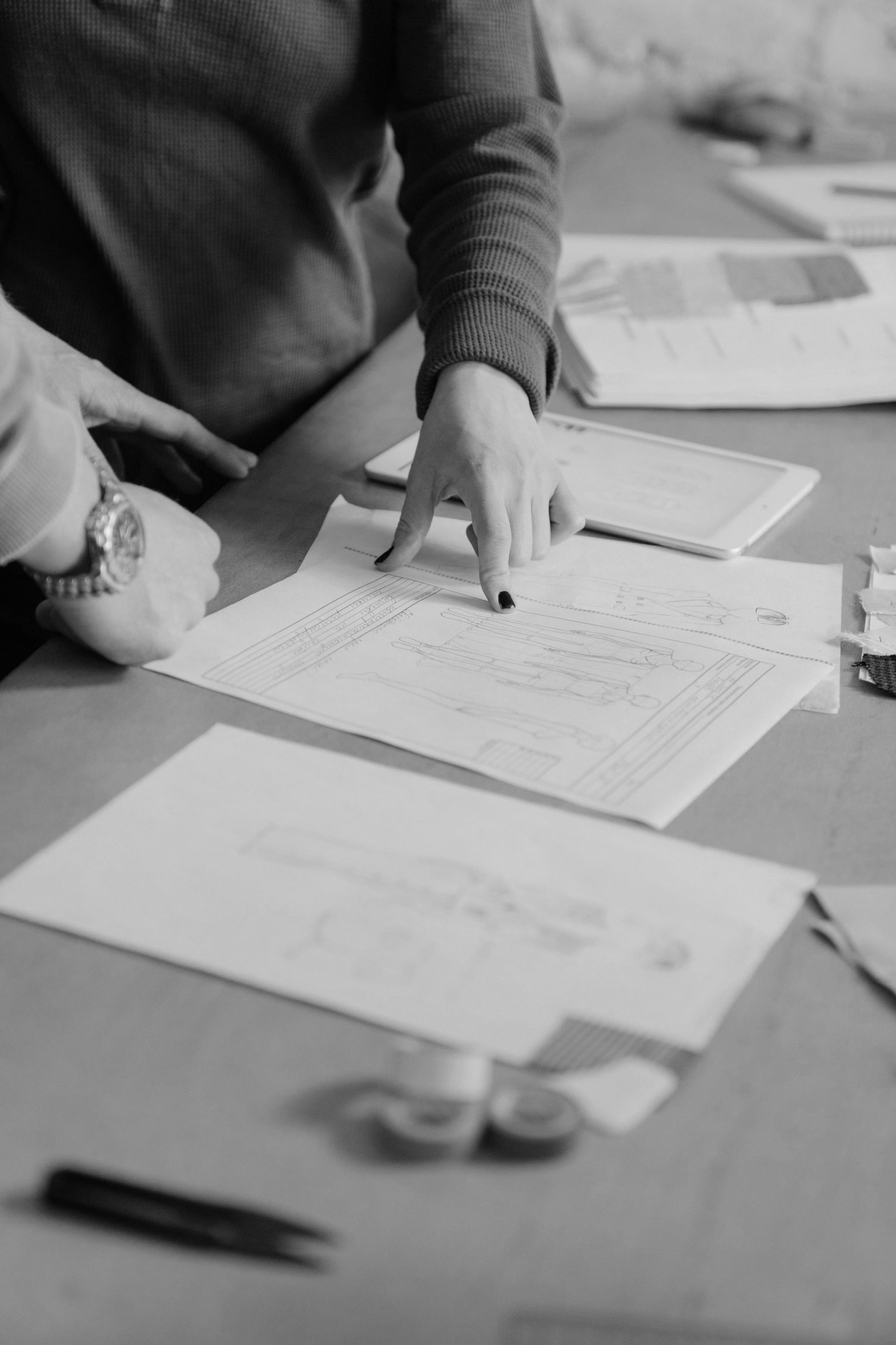 İki kişinin üzerinde moda tasarım çizimleri ve tablet bulunan masada çalışması.